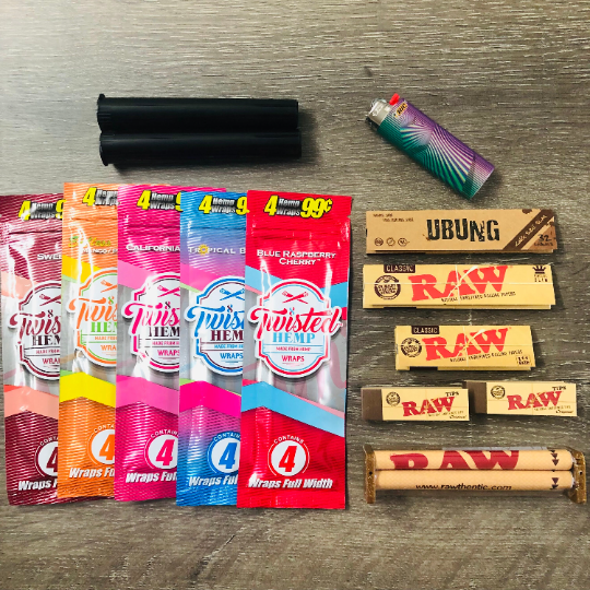 Photo of the contents are laid out on a table displaying five hemp wrap, rolling papers, rolling machine, joint tubes, RAW tips, and a lighter.