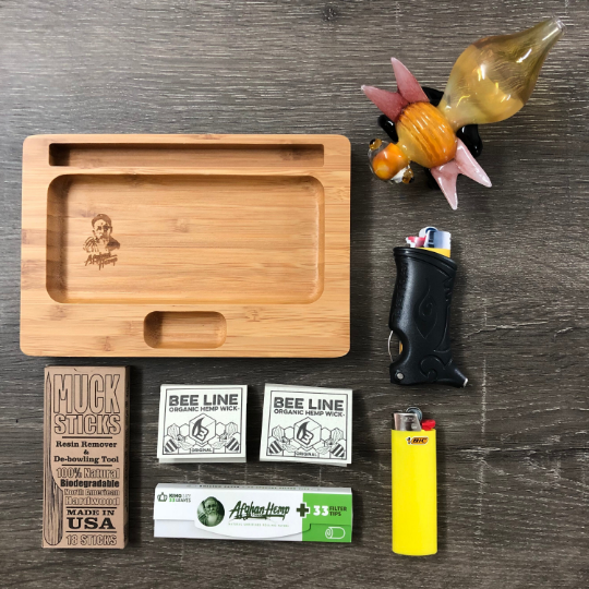 Photo displays the contents laid out on a table that includes a rolling tray, beeline hemp wick, toker poker smoking tool, rolling papers, muck sticks, and a lighter.