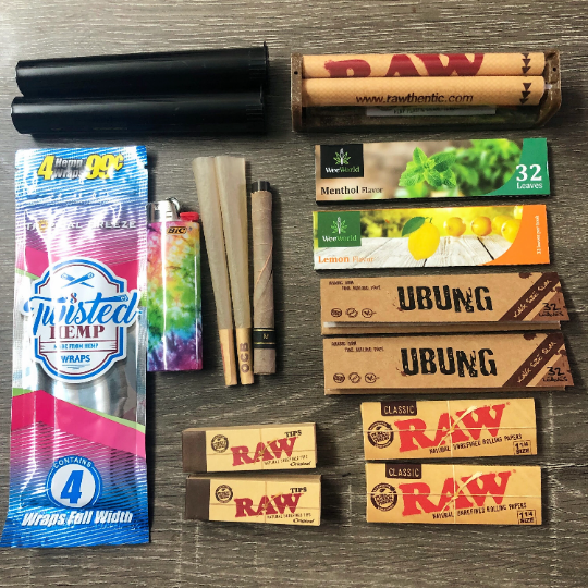 Photo of the contents are laid out on a table displaying a hemp wrap, rolling papers, rolling machine, individual cones, joint tubes, RAW tips, and a lighter.