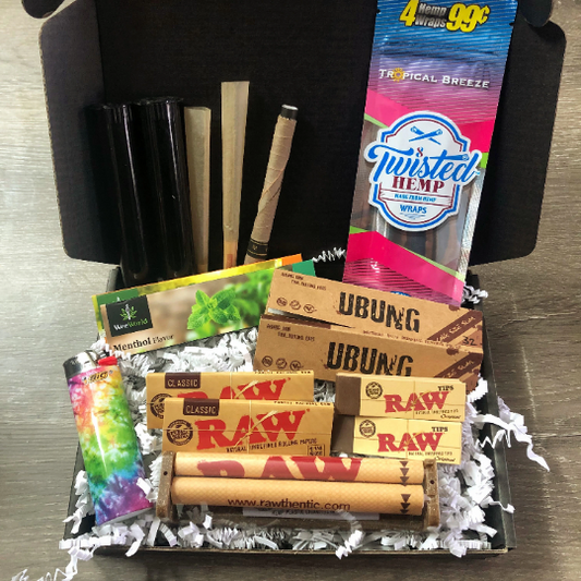 Photo of a black gift box with white shredded paper. Contents in the gift box includes a hemp wrap, rolling papers, rolling machine, individual cones, joint tubes, RAW tips, and a lighter.