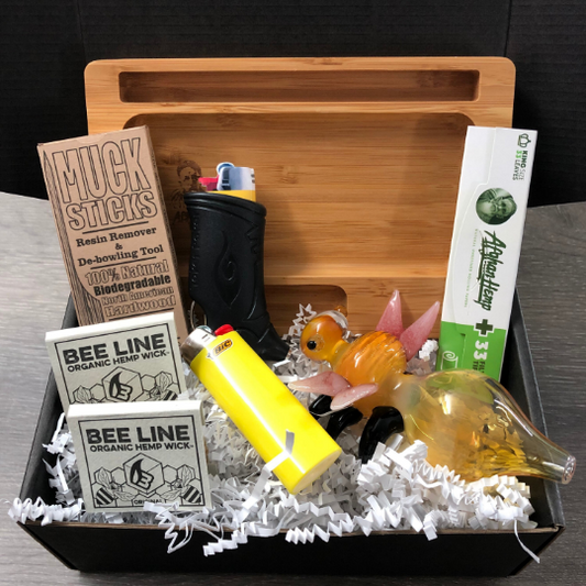 Photo of a black gift box with white shredded paper. Contents in the gift box includes a rolling tray, beeline hemp wick, toker poker smoking tool, rolling papers, muck sticks, and a lighter.